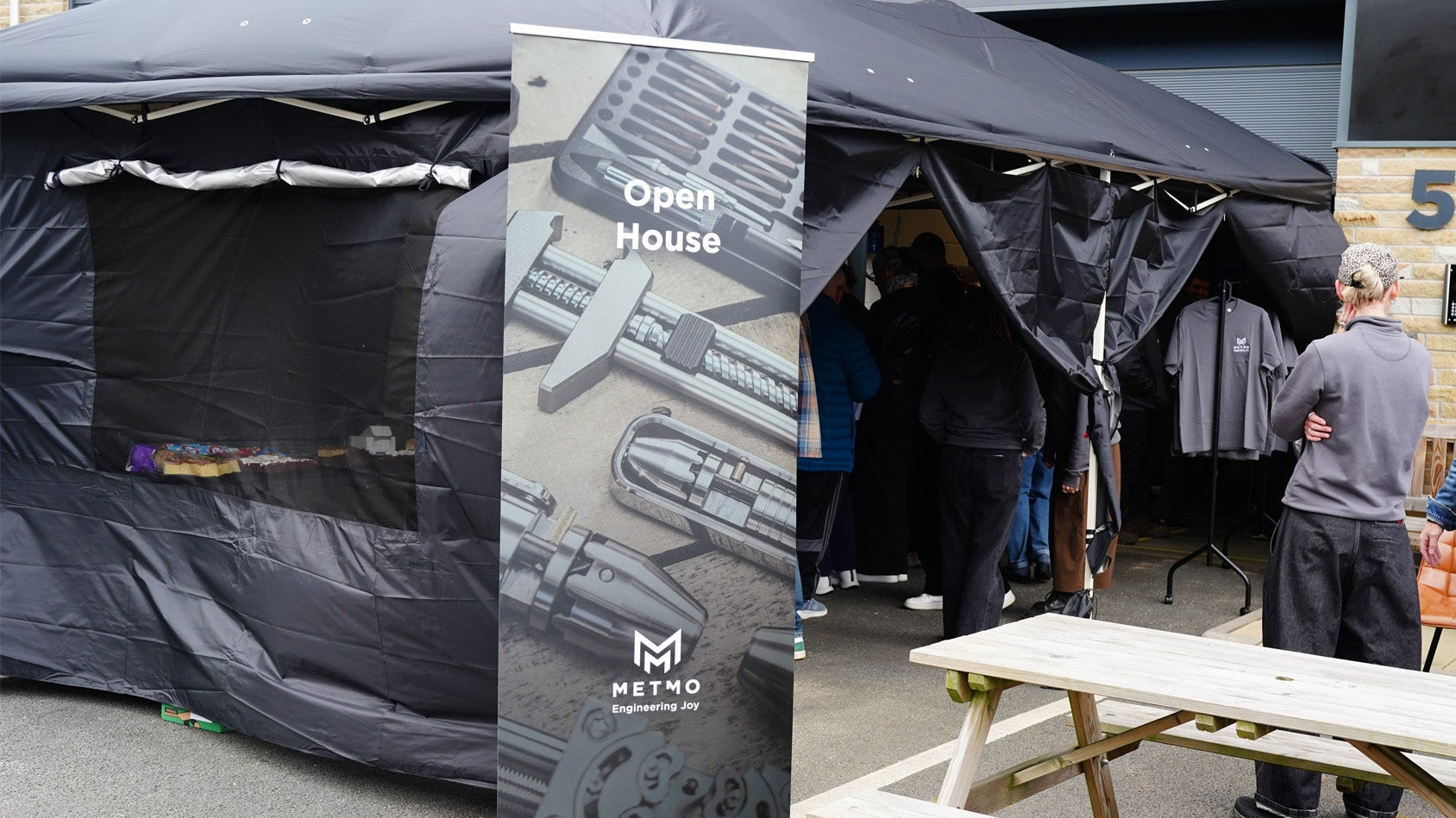 Outdoor event setup with a black canopy tent, a vertical METNO roll-up banner, people standing and entering the tent, and a wooden picnic table in the foreground.
