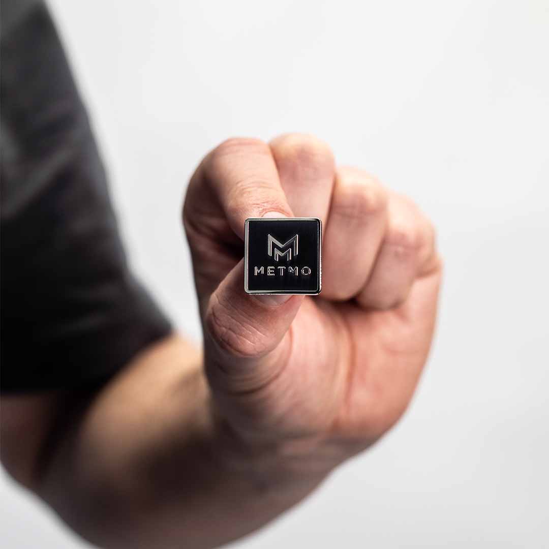 Hand holding a black square pin badge with 'METMO' branding against a light background