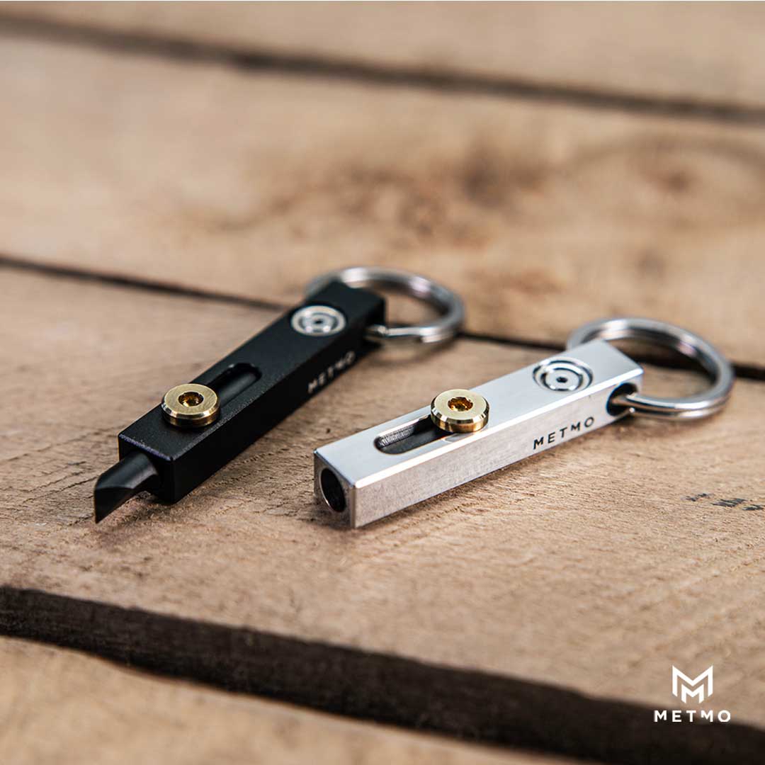 Two Edge keychains, one black and one silver with gold accents, on a wooden surface with 'METMO' branding.
