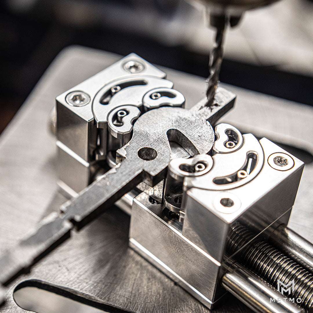 Close-up of a metal key in a precision fractal vice, with a drill bit positioned above it on a brushed metal work surface.