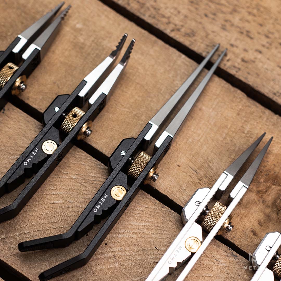 Close-up of several metal precision tweezers arranged on a rustic wooden surface. The tweezers are shown in black and silver finishes, with one pair featuring gold-toned adjustment screws.