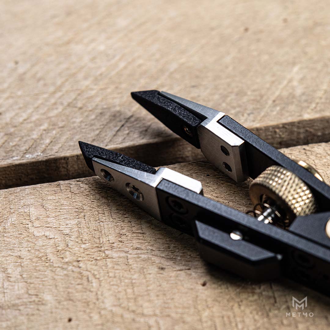 Close-up of a black and silver metal tool with brass-colored screws and a textured gold knob, resting on a rustic wooden surface.