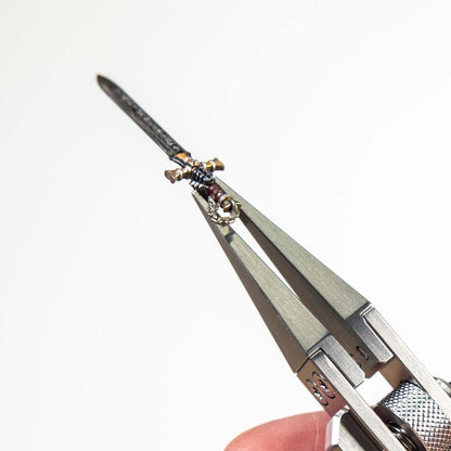 A miniature metal sword accessory held in MetMo Precision Grip against a plain white background, with a fingertip visible at the bottom.