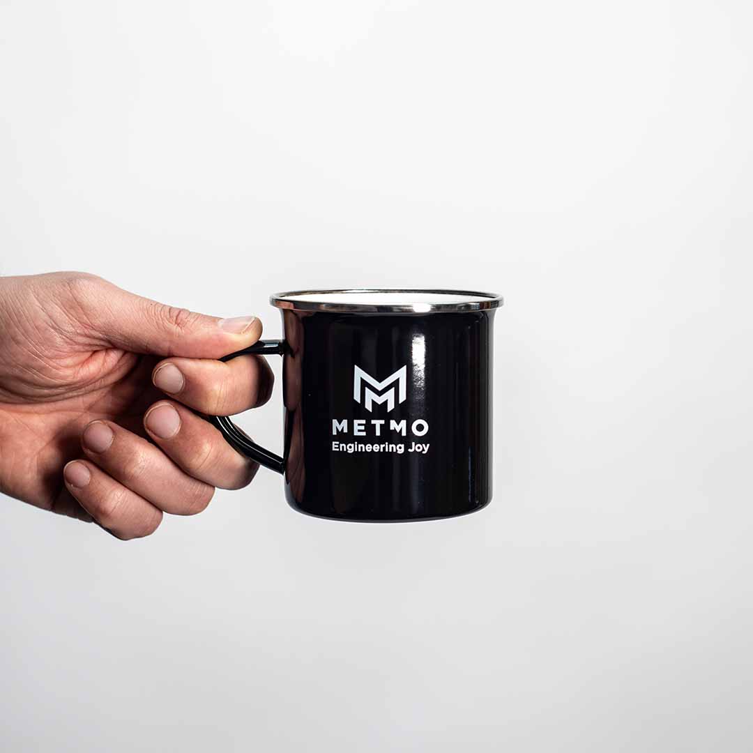 Black mug with 'METMO Engineering Joy' logo held by a hand on a white background