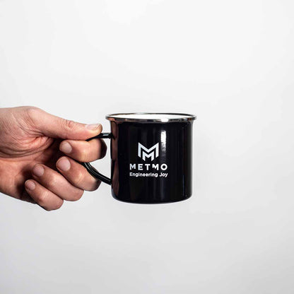 Black mug with 'METMO Engineering Joy' logo held by a hand on a white background
