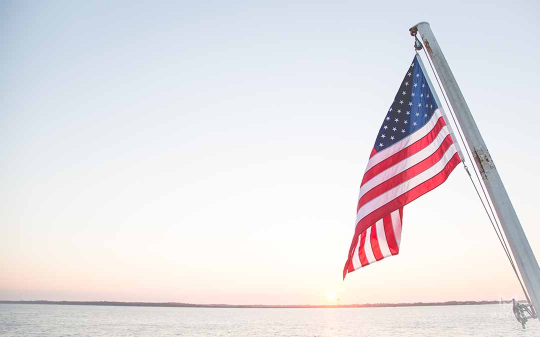 American flag hanging from a pole with a muted sea and sunset background
