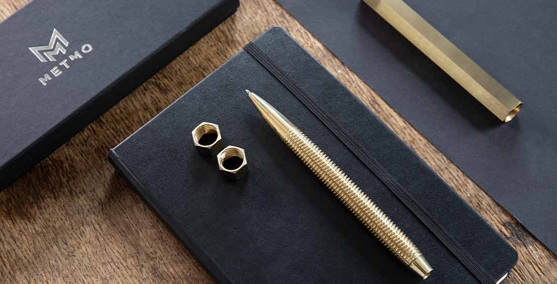 Hero image of brass pen with nuts, metal case, and notebook