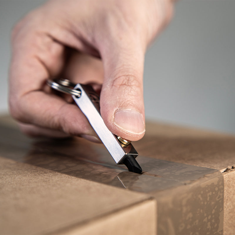 Hand using a MetMo Edge to open a cardboard box on a neutral background