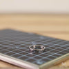A close-up of a silver geometric ring held in a MetMo Precision Grip, resting on a black gridded cutting mat with a blurred wooden background.